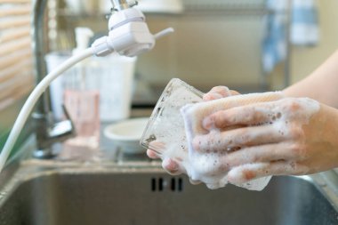 Temizlikçi kadın lavaboda herkesin hijyen ve virüsten korunması için kullanılmış içki bardağını yıkıyor. Temizlikçi, bardakları ve bulaşıkları yıkamak için deterjan ve sünger kullanıyor..