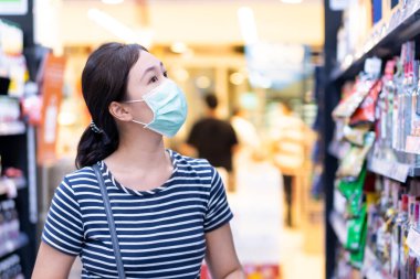 Coronavirus ya da COVID-19 salgını sırasında markette yürüyen ve raflarda mal arayan genç bir Asyalı kadın. Süpermarkette su arayan koruyucu maskeli bir kadın.