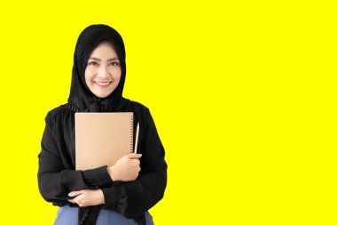 20 'li yaşlardaki güzel Asyalı Müslüman kadın portresi. Renkli konsept. Zeki Müslüman öğrenci elinde bir defter ve kalem tutarak sarı renkli duvarda fotoğraf için poz veriyor..