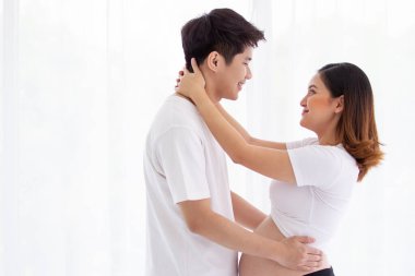 Asyalı güzel karı koca portresi, karısı hamile. Koca şefkatle karısını yatakta kucaklıyor ve öpüyor. Adam hamile karısını kucaklıyor ve ellerini kalp şeklinde yapıyor.