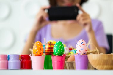 Asyalı genç ve güzel bir kadın akıllı telefon kullanarak renkli bir Paskalya yumurtasının fotoğrafını çekmeyi seviyor. Kovada renkli Paskalya yumurtası. Paskalya yumurtası festivali konseptinin sembolü..