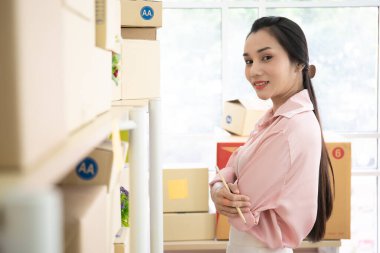 Çevrimiçi Genç Asyalı kadın tüccar güvenli bir şekilde parsel rafının önünde duruyor. Rafta sevkiyat için hazırlanmış bir sürü paket var. Ticaret ticareti konsepti.