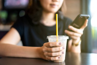 Asya kökenli genç bir kadın kahvecide rahatlıyor ve buzlu kahve içiyor. Kafe ve restoranda kahve içerken mesaj göndermek için akıllı telefon kullanan bir kadın..
