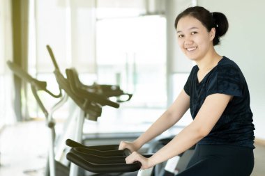Asyalı şişman kadın kapalı spor salonunda bisiklet antrenmanı yapıyor. Bisiklet makinesiyle kişisel egzersiz yapan bir kadın. Kadın vücudunda bakım ve refah. Spor salonunda spor yapan neşeli şişman kadın..