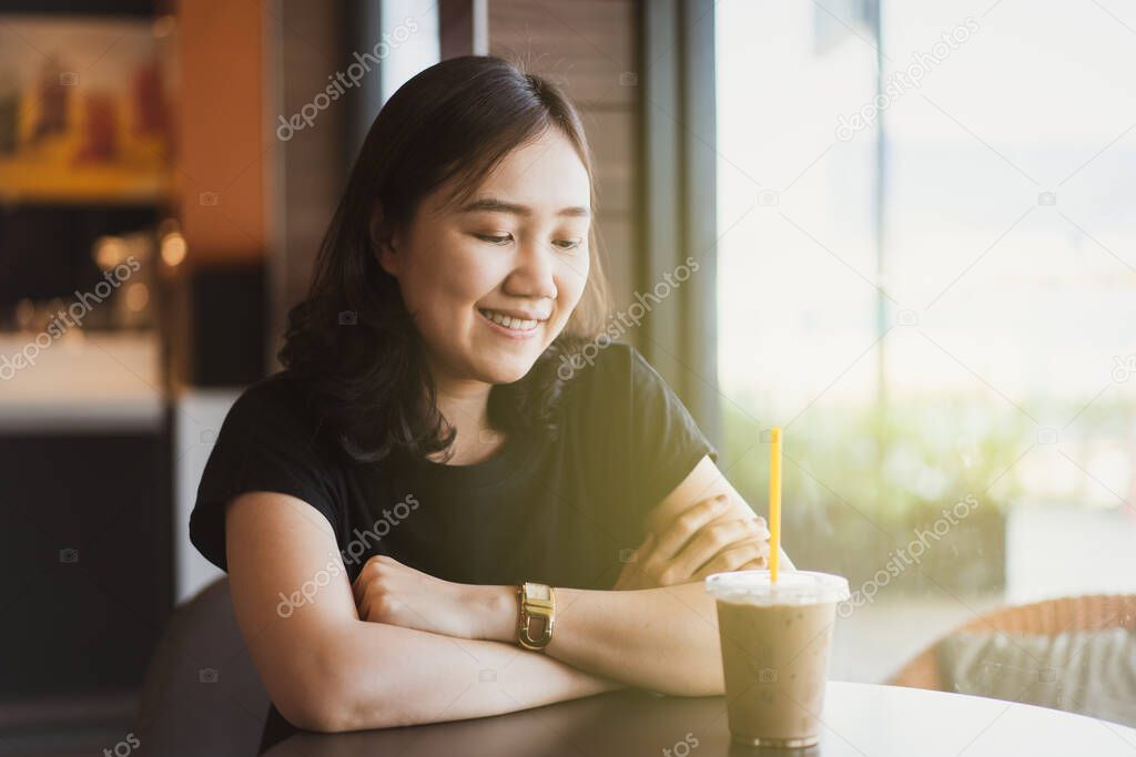 Beautiful young Asian woman portrait in coffee shop, woman enjoy drinking iced dark chocolate in a cup on the table. Woman satisfied drinking iced dark chocolate at modern co-working space and cafe.