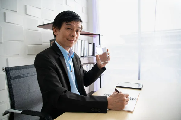 アジアの高齢者のビジネスマンは彼のオフィスに滞在し 高齢者の従業員は仕事中にリフレッシュするために水を飲む 高齢者の健康とワークライフバランスの概念 ビジネスマンはリラックスして酒を飲んで — ストック写真