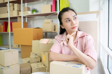 Düşünceli Asyalı kadın oturma odasında bir sürü paket kutusuyla oturuyor. Elinde kalem tutan ve online satış işini düşünen bir kadın. Lojistik ve servis konsepti.