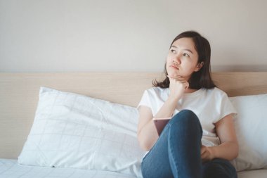 Asyalı güzel kadın dinleniyor ve yatak odasındaki fotokopi odasının üzerinde tablet kullanıyor. Neşeli kadın yataktan kalktıktan sonra tablet kullanarak internete ya da sosyal medyaya göz atıyor. Kadın düşüncesi.