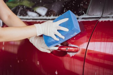 Genç Asyalı bir kadın, yumuşak bir sünger ve sıvı sabun kullanarak bir arabayı yakın mesafeden yıkıyor. Kadın garajda modern kırmızı arabayı yıkayıp fotokopi aletiyle kapatıyor..