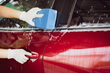 Genç Asyalı bir kadın, yumuşak bir sünger ve sıvı sabun kullanarak bir arabayı yakın mesafeden yıkıyor. Kadın garajda modern kırmızı arabayı yıkayıp fotokopi aletiyle kapatıyor..