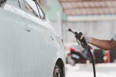 Garajda çalışan adam yüksek basınçlı köpük enjektörüyle kirli beyaz bir arabaya püskürtüyor. Yağmur mevsiminde araba yıkama ve araba bakım konsepti. El emeği kirli bir arabaya sıvı sabun püskürtür..
