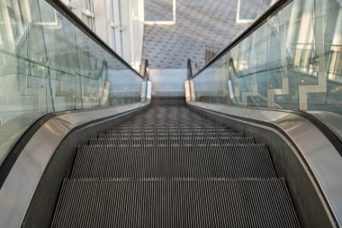 Downward view of empty escalator inside modern building, metallic steps and glass handrails in clean architectural environment.