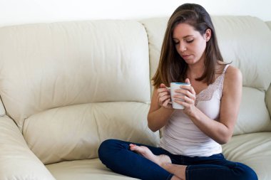 Esmer kız ve onun evinde oturma odasında kanepede kahve, çay veya infüzyon tutan yalnız, dalgın genç kadın