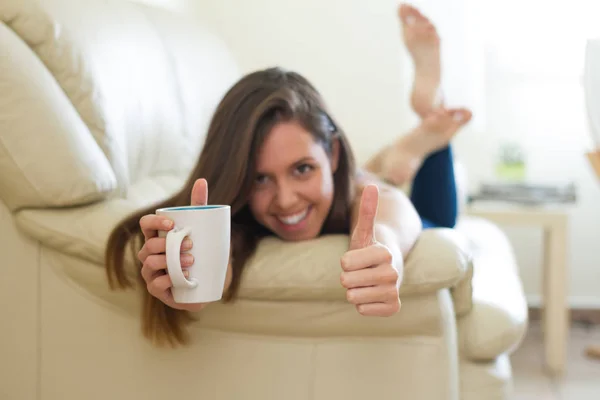 Young brunette girl with fingers in okey position, thumbs up, approving situation, happy, empowered while taking a cup of hot coffee in the living room of her house