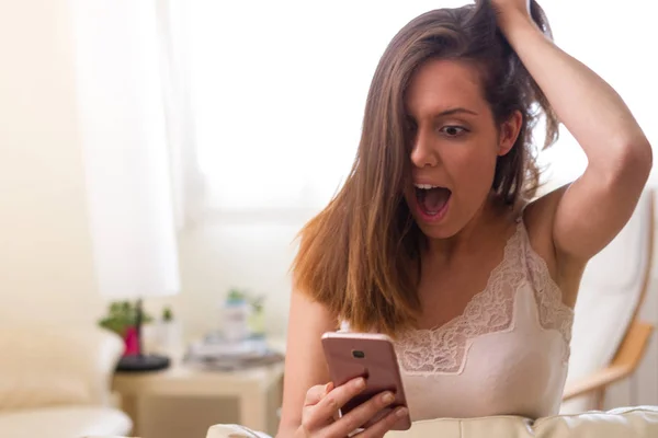 European brunette woman with surprised brown eyes pulling her hair while observing her mobile phone which has received a message that provokes nervousness impression and surprise