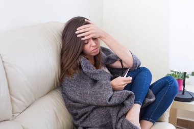 Esmer beyaz olan kadın baş ağrısı, karın ağrısı, hasta, bir battaniye ile kanepede oturan bir termometre ile ateş ve ağrı, hastalık, soğuk, ıssızlık, üzüntü ve çaresizlik gösterilen kafasına el