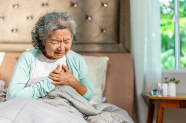 Asyalı olgun bir kadın yatak odasındaki yatakta kalp krizi geçiriyor. Yaşlı kadın göğüs bölgesinde ağrı ve nefes darlığı çekiyor. Acil sağlık ve sağlık sigortası kavramı.