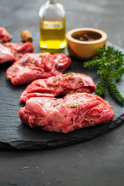 Raw pork cheeks meat on a dark gray surface with rosemary and spices. Food background with copy space. Vertical. Space for text.