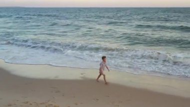 Mutlu çocuk sahilde koşuyor. Günbatımı ılık ışık, altın kum, okyanus kıyısında komik çocuk oyunu. Mavi dalgalarla kumsalda yürüyen küçük güzel çocuğa hava manzarası. 