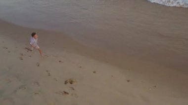 Mutlu çocuk sahilde koşuyor. Günbatımı ılık ışık, altın kum, okyanus kıyısında komik çocuk oyunu. Mavi dalgalarla kumsalda yürüyen küçük güzel çocuğa hava manzarası. 