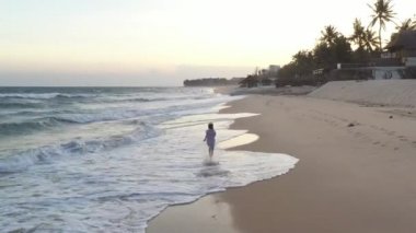 Mutlu çocuk sahilde koşuyor. Günbatımı ılık ışık, altın kum, okyanus kıyısında komik çocuk oyunu. Mavi dalgalarla kumsalda yürüyen küçük güzel çocuğa hava manzarası.  