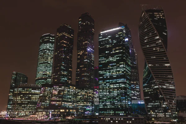 Horizontal shot of big city high glass buildings of different shape, with lighted windows at night, on dark brown sky background. One of the buildings has interesting modern spiral shape. — Stock Photo, Image