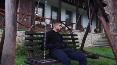 Genç bir adam büyük bir ev sundurma yakınındaki bir salıncak üzerinde oturur ve sis ormanda bakar