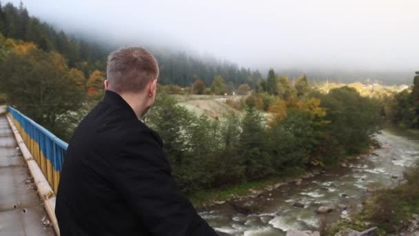 一个年轻的家伙从桥到一条山河 在喀尔巴阡山脉的雾中看到一座山 — 图库视频影像