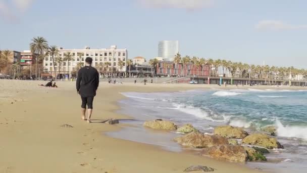 Un tipo con ropa caminando en la orilla de un mar frío y ondulado en Barcelona — Vídeos de Stock