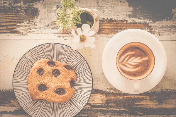 big chocolate chip cookie with a cup of cappunico with latte art on top and a small vase of flowers