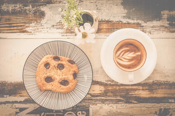 big chocolate chip cookie with a cup of cappunico with latte art on top and a small vase of flowers