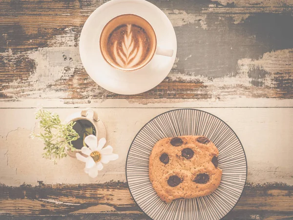 big chocolate chip cookie with a cup of cappunico with latte art on top and a small vase of flowers