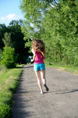 Genç kadın atlet. Kadın yeşil ağaçlarıyla çevrili toprak yol üzerinde çalışır. Arkadan, fotoğraf yüz olmadan.