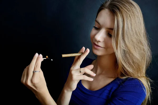 Young female lighting up cigarillo with matches. Woman in blue dress with black background.