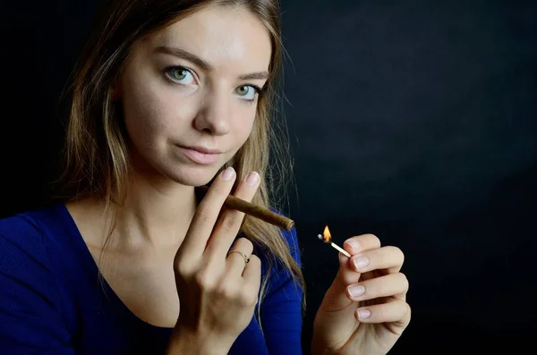 Young female lighting up cigarillo with matches. Woman in blue dress with black background.