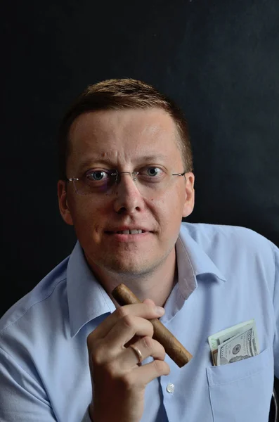 Male model, 35 years old. Man with glasses, wearing light blue shirt, holding cigar in his hand. US Dollars banknotes in shirt's pocket.