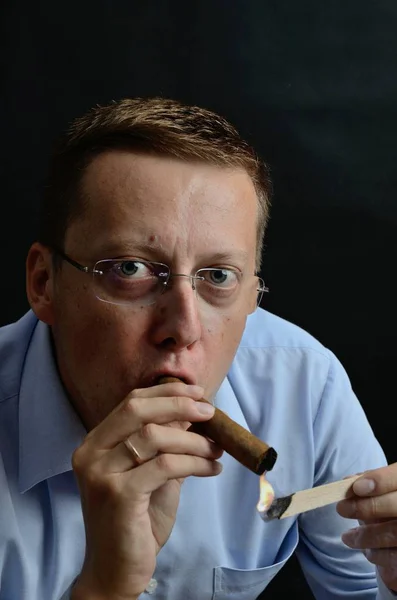 Male model, 35 years old. Man with glasses, wearing light blue shirt, lighting up a cigar.