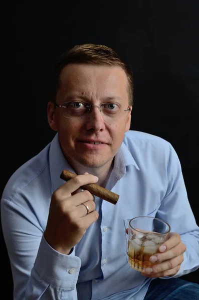 Male model, 35 years old. Man with glasses, wearing light blue shirt, holding cigar and glass of whiskey.