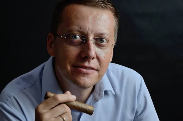 Male model, 35 years old. Man with glasses, wearing light blue shirt, holding cigar in his hand.