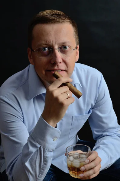 Male model, 35 years old. Man with glasses, wearing light blue shirt, holding cigar and glass of whiskey.