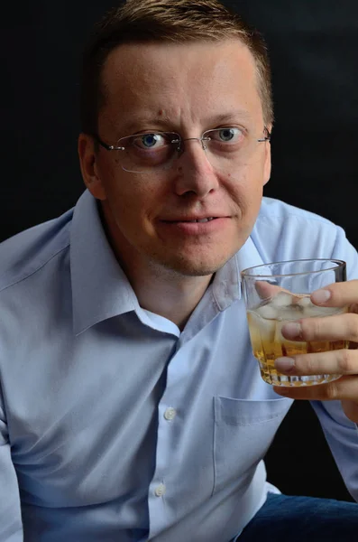 Male model, 35 years old. Man with glasses, wearing light blue shirt, holding glass of whiskey with ice.