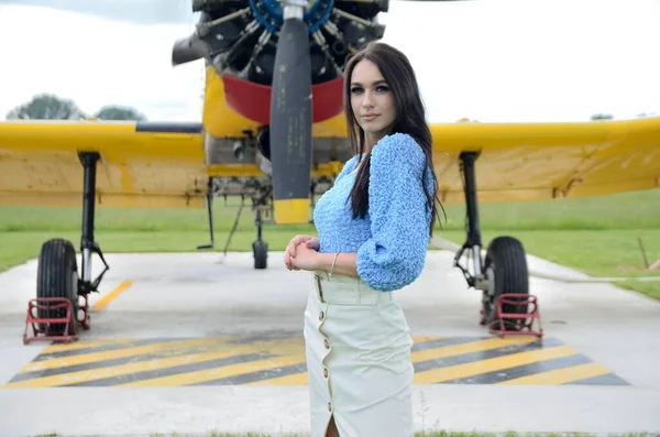 飛行機のある若いエレガントな女性 青いトップと白いスカートの女性がポーランドから黄色の飛行機の前に立つ — ストック写真