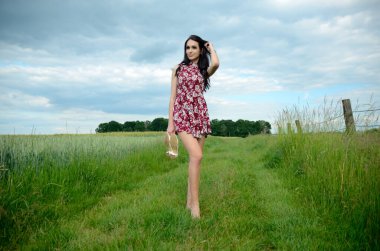 Beautiful woman holding her shoes in hands, barefoot on dirt road in countryside. Young Polish woman surrounded by green grass. clipart
