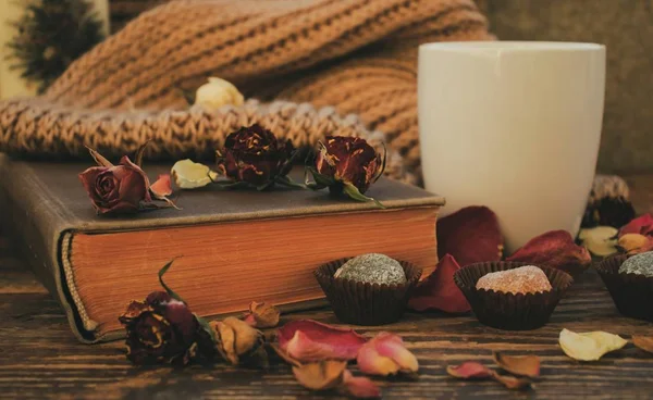 A mug of tea and a book surrounded by dry flowers and candy.