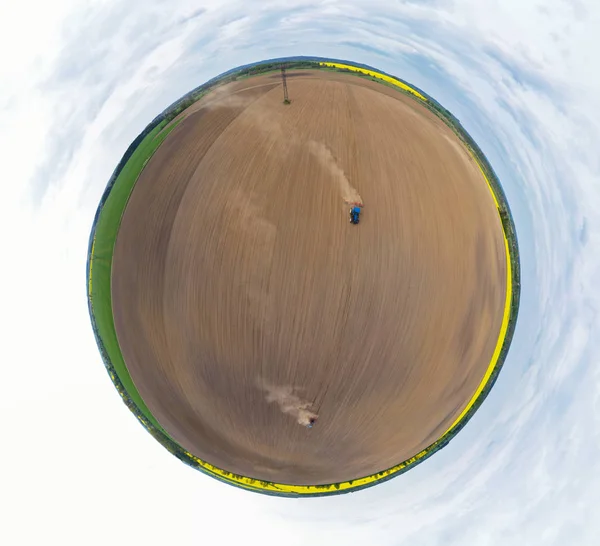 Vista panorámica aérea de 360 grados sobre tractor azul tirando de un arado, preparando un suelo para la siembra de semillas, tractor haciendo nube de tierra. Monumento en prado y campo de colza —  Fotos de Stock