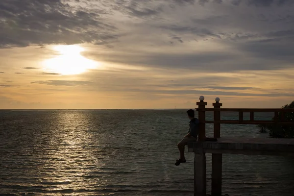 Silhueta de um homem sentado e olhando para o mar sozinho na borda da ponte com belo céu por do sol. Royal Island, Indonésia — Fotografia de Stock