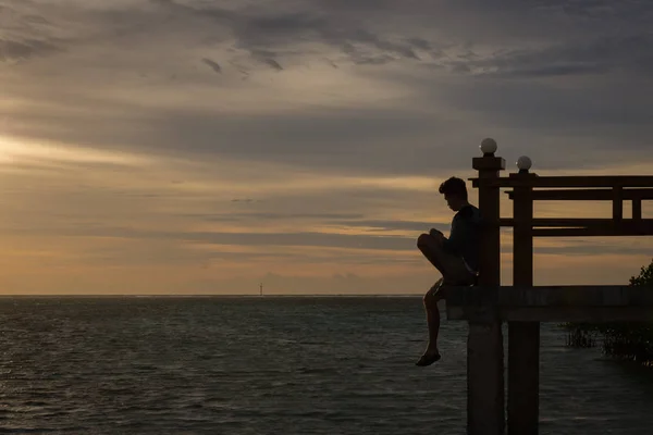 Silhouette of a man sitting and looking at the sea alone on the edge of the bridge with beautiful sunset sky. Royal Island, Indonesia