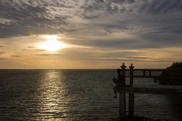 Silhueta de um homem sentado e olhando para o mar sozinho na borda da ponte com belo céu por do sol. Royal Island, Indonésia — Fotografia de Stock