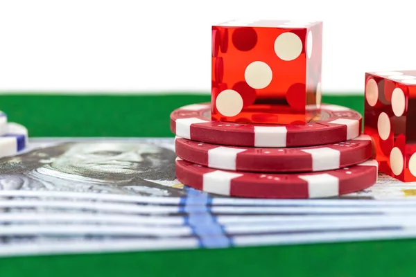 Poker chips and dices lie next to blurry banknotes with a portrait of Benjamin Franklin, side view on a green background / concept of a game in a casino