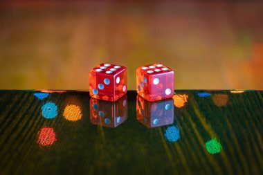 Cilalanmış ahşap bir masada iki kırmızı cam zar, arka planda bulanık, çok renkli ışıkların yansıması. Kutlama zaferi konsepti. Kopyalama alanı olan altı ve altı.